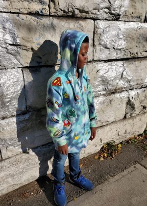 A boy in a superman jacket standing on the sidewalk.