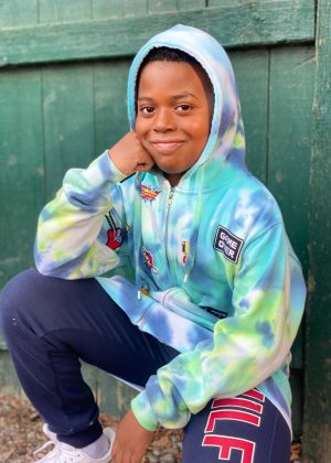 A young boy in a tie dye hoodie sitting on the ground.